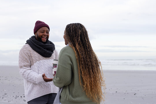 African couple laughing, chatting on sandy shore holding patterned cup and tote, wearing knits