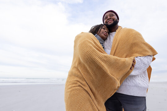 Couple standing wrapped in mustard-yellow blanket on sandy beach, man wearing beanie and sweater
