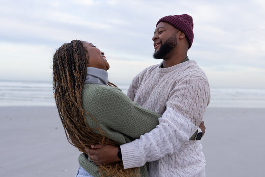 African American couple embracing, laughing on sandy shore, wearing knit sweaters, beanie, scarf