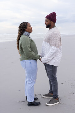 African couple holding hands on wet sandy beach near shoreline, wearing knit sweaters and beanie