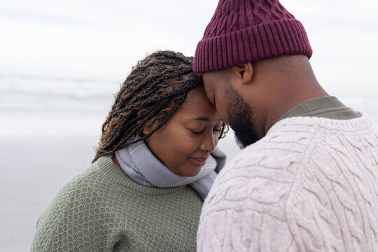 African adult couple standing touching foreheads on sandy shore wearing knit sweaters and beanie