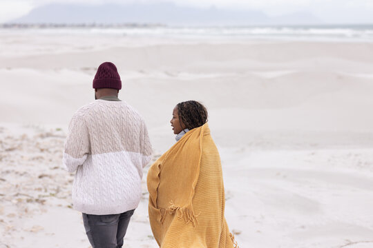 African American couple walking along windswept beach, wearing mustard blanket and cream sweater