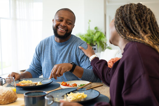African American couple smiling, eating breakfast at dining table near window with plates, juice