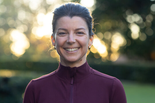 Woman smiling and basking in golden light in park with maroon zip-up and stud earrings