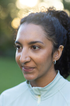 African woman looking left outdoors in sunlit park, wearing zip-up jacket, nose ring, ear cuff