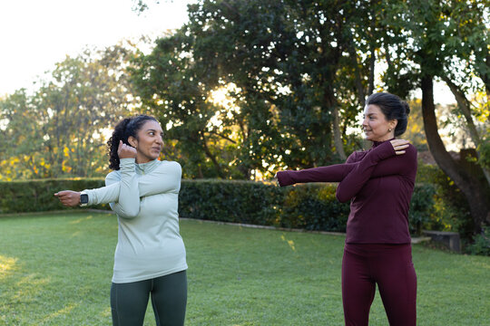 Diverse female friends stretching on backyard grass, wearing sportswear with smartwatch