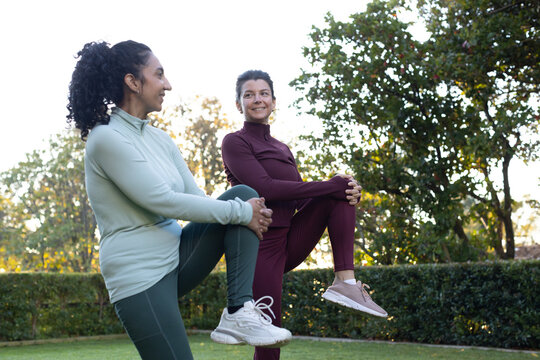 Diverse adult female friends holding knees and balancing on lawn, in long-sleeve tops and sneakers