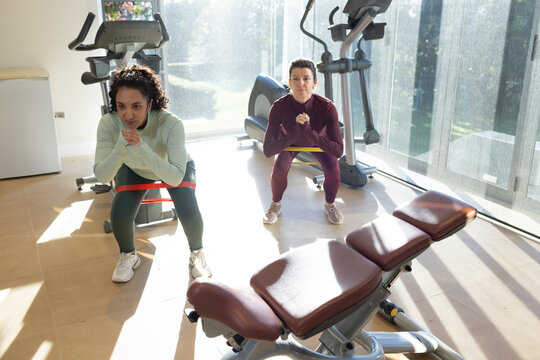 Two women in athletic wear performing resisted squats with loop bands at home gym bench