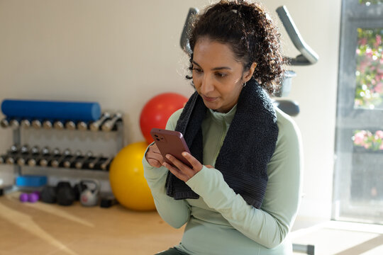 African American woman sitting on bench in home gym, holding phone, wearing green top and towel