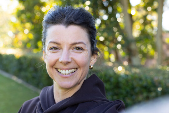 Woman smiling, wearing dark coat with high collar in garden with sunlight showing gold hoop earring