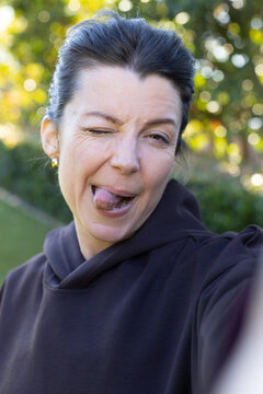 Woman winking and sticking out tongue, holding phone in garden wearing dark hoodie and gold studs
