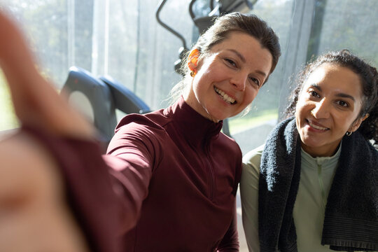 Two female friends smiling and posing for selfie in gym wearing maroon green tops by treadmills