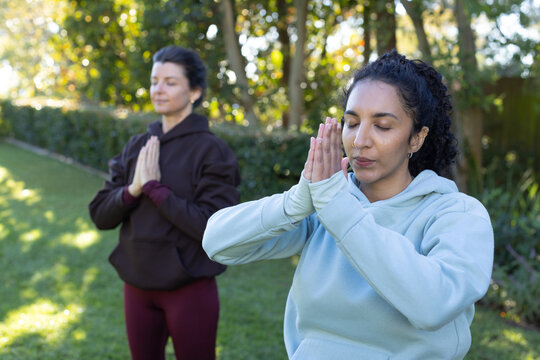 Diverse female friends practicing prayer pose on grassy backyard lawn wearing hoodies and leggings