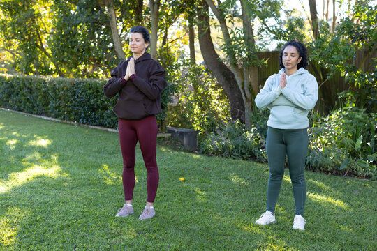 Diverse female friends practicing breathing exercise on backyard lawn wearing hoodies and sneakers