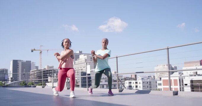 Two women starting workout, walking then lunging in sync on rooftop near dumbbells in workout gear