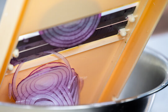 A red onion is being thinly sliced on a plastic mandoline slicer over a metal bowl, creating perfect concentric rings.