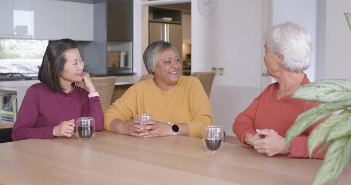 Diverse women chatting at home table with mugs, right woman starting story, center gasping smiling