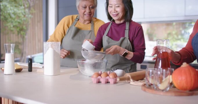 Diverse women helping as center tapping sifter sifting flour into bowl for dough at kitchen island