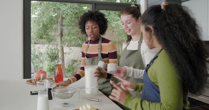 Diverse female friends agreeing recipe, scooping flour from jar at counter, in aprons to bake