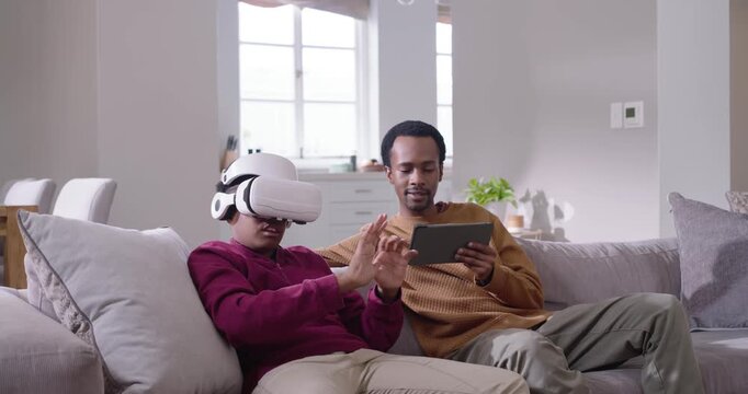 African American men putting on white VR headset, gesturing while tablet-user guiding hands at home