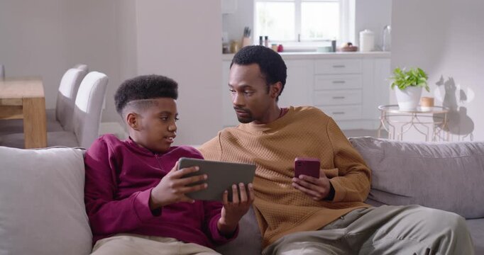 African American father and teen leaning after tablet shift sharing phone screen, laughing on sofa