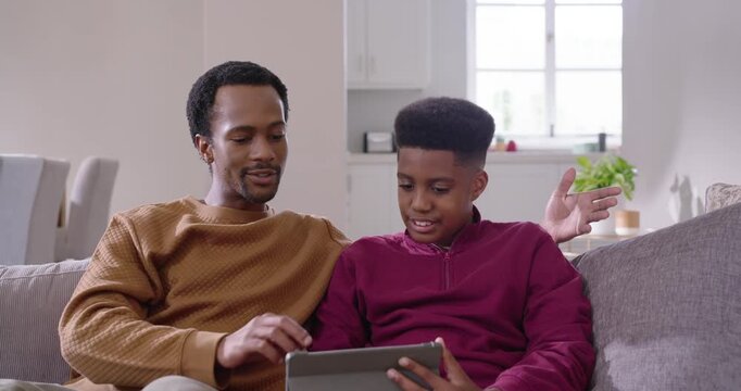African American dad and teenage son sitting on sofa, dad reaching tablet, helping, fist bumping