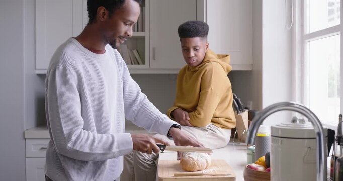 African American dad putting loaf at kitchen counter slicing teenage son reaching jars making snack