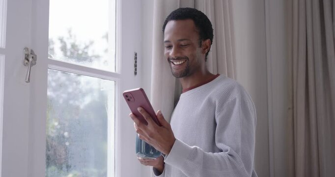 African American man reading pink phone text, smiling, laughing, sipping blue mug at home by window