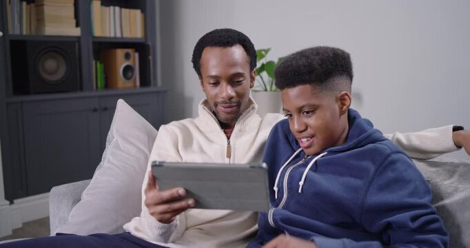 African American dad and teen son sitting on sofa, teen tapping tablet, father adjusting, smiling