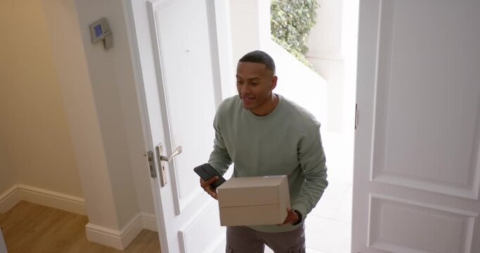 African American adult male checking phone while carrying box, stepping through front doorway