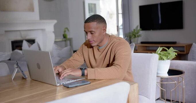 African American man in tan sweater typing on laptop at dining table, responding to work emails