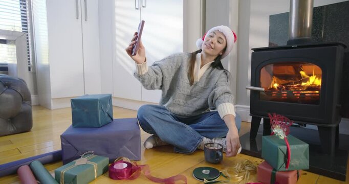 Woman wearing Santa hat at home by stove, receiving call, showing wrapped gift to phone, sharing