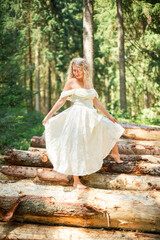 Fototapeta premium Blonde woman in a flowing white off-shoulder dress carefully walks barefoot across fallen tree trunks while holding her skirt in a sunlit forest. Romantic outdoor scene