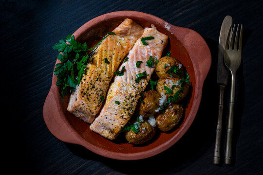 Baked salmon fillet with roasted potatoes in clay plate, closeup. Delicious baked salmon fish fillet served with small roasted potatoes and fresh herbs in rustic clay dish on dark wooden background
