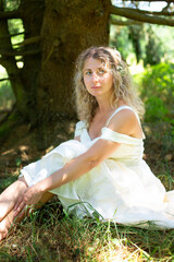 Fototapeta premium Blonde woman in a long white off-shoulder dress sits on the forest floor beneath the spreading branches of a large tree