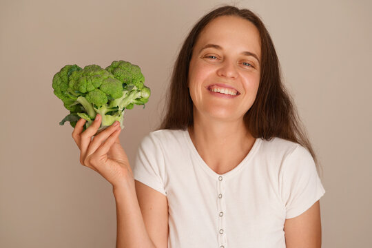 A woman stands with a bunch of broccoli in her hand. She looks happy and is smiling. The setting has a plain background and it is daytime
