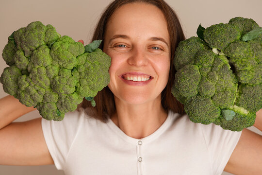 A woman stands indoors in good light. She smiles while holding two large broccoli heads by her face. Green vegetables are fresh and large
