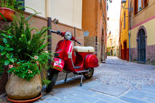Vintage red scooter parked beside a potted plant on a narrow cobblestone street in the historic center of Bosa, Sardinia, Italy