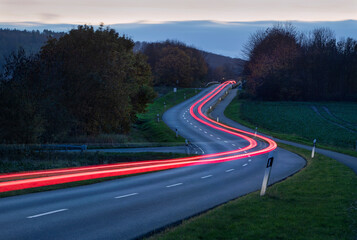 Lichtstreifen eines vorbeifahrenden Fahrzeugs auf einer kurvigen Landstraße in der Dämmerung. © thomaslerchphoto