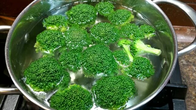 A metal pot of boiling water containing bright green broccoli florets sits on the stove, conveying the impression of a fresh and healthy cooking process.