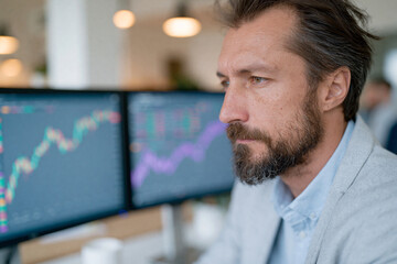 Fototapeta premium Focused man studies financial charts on dual monitors. His serious gaze shows concentration and intent. The office lighting is soft, ambient, and professional. Ideal for business, finance