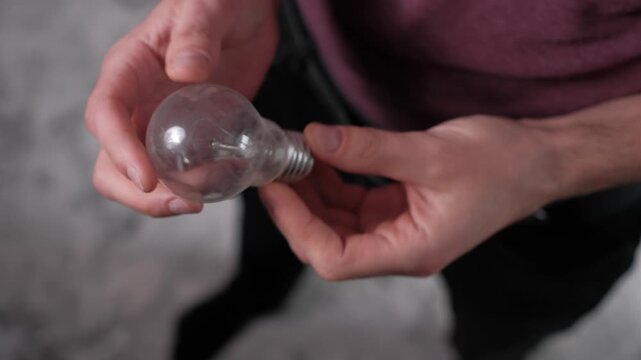 Overhead close-up of male hands rotating a clear incandescent bulb to reveal the inner filament. The glass texture and screw base symbolize outdated lighting technology.