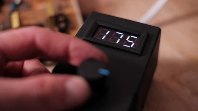 Fingers rotate a control knob to decrease the numerical voltage value on a bright LED screen. The action illustrates precise electrical adjustment and laboratory calibration.