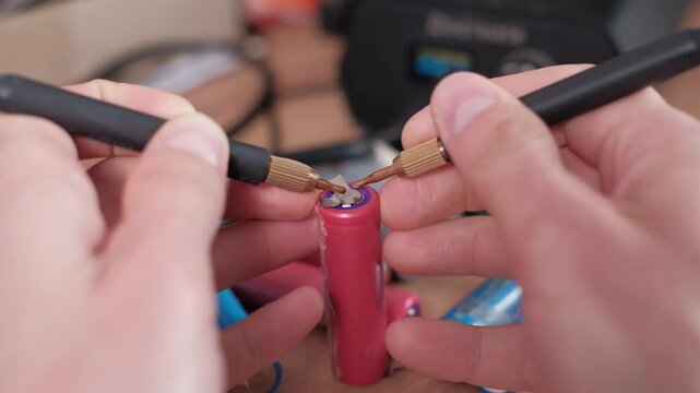 Detailed view of hands using a spot welder to attach a nickel tab onto a pink lithium-ion battery. The workshop setting suggests custom electronics repair and renewable energy work.