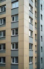 Fototapeta premium Apartment building exterior detail showing multiple floors with beige and grey facades. White framed windows are arranged in repeating pattern. Side view reveals corner of multistorey structure.