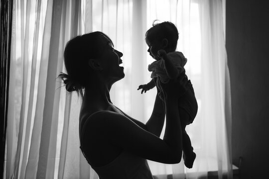 ilhouette of a mother lifting her baby up in front of a window, cozy home atmosphere. Black and white photo