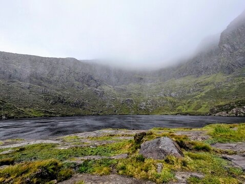 Moody and atmospheric view of a dark mountain lake surrounded by steep rocky cliffs covered in thick fog at Conor Pass, Ireland.