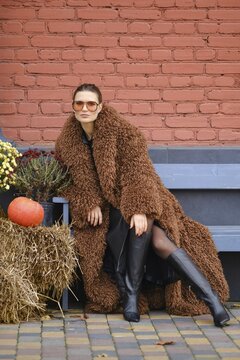 A woman wearing a brown fuzzy coat and knee-high black boots poses confidently outdoors. She is surrounded by autumn decorations, including a pumpkin, haystack and flowers, set against a brick wall
