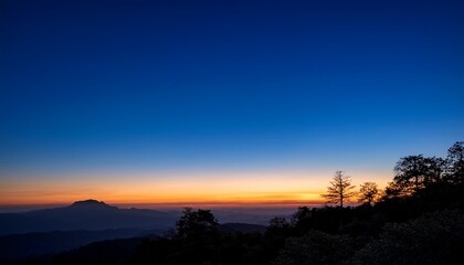 Fototapeta premium Silhouetted Treetops And Distant Mountains Lie Under A Vast Clear Sky Transitioning From The Deep Blue Of Twilight To The Warm Orange Glow Of Dawn On The Horizon