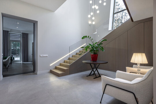 A bright entryway is shown with a staircase, a potted plant on a table, and a lamp. Sunlight enters through large windows, highlighting the design of the space.